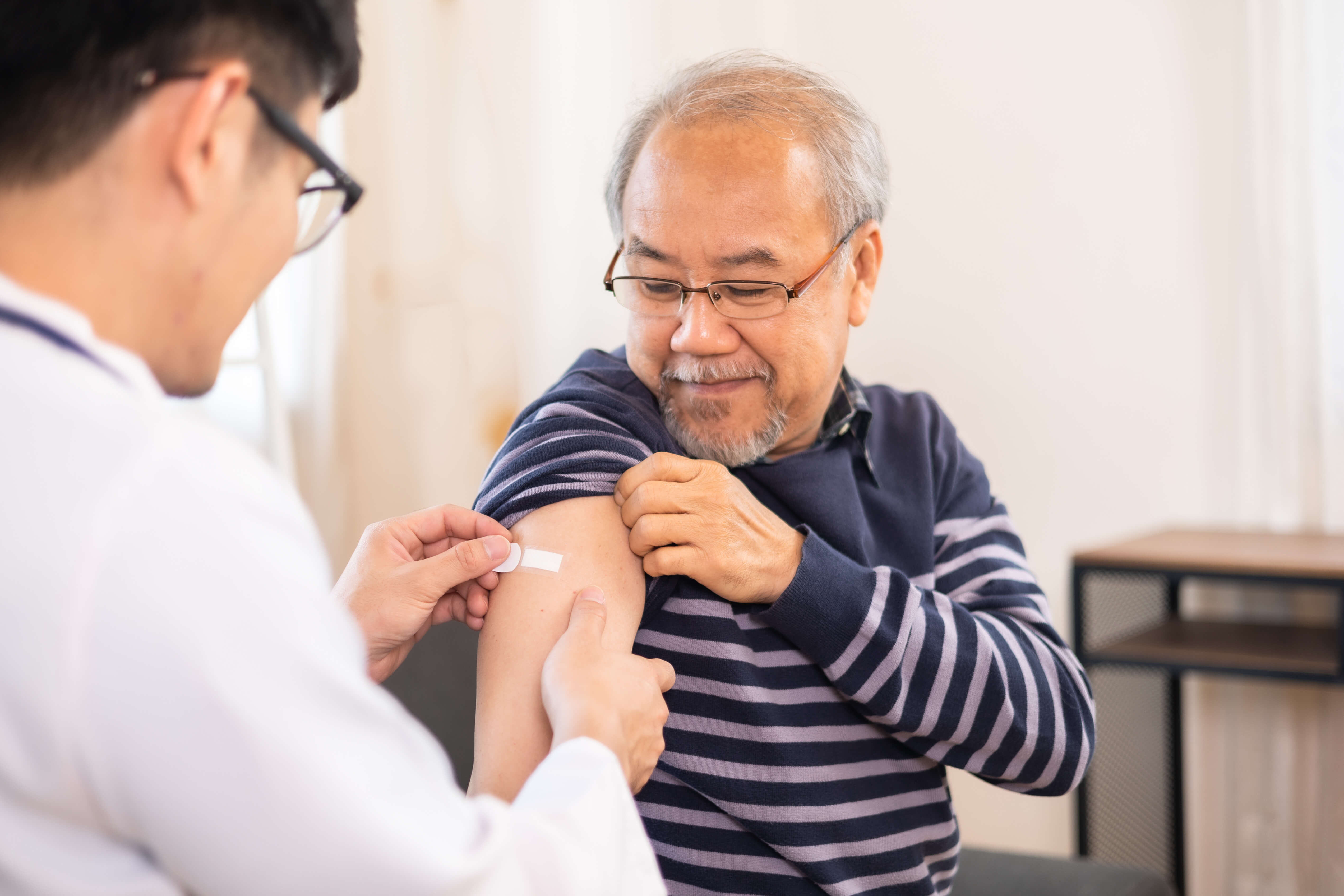 An older asian male getting a flu vaccine from a doctor. He is smiling down at his arm when the doctor is putting on a bandaid.