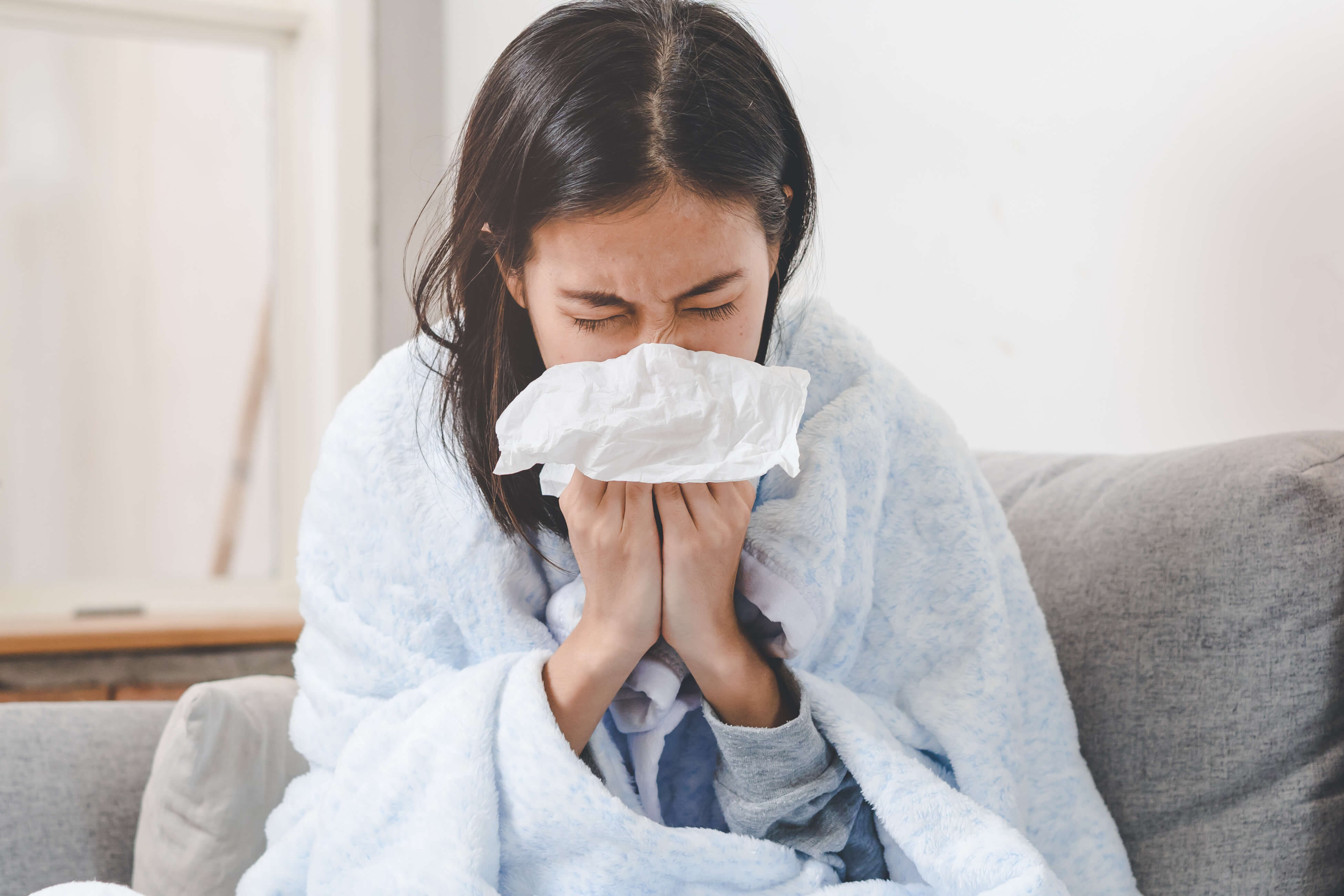 An asian woman at home with a blanket around her arms, blowing her nose.